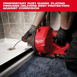 The Milwaukee Fuel drain cleaner is being used by a person wearing black gloves. The tool's red casing is visible against a background of a tiled bathroom and a blue rug. Text above describes the product's rust guard plating process that offers superior corrosion protection.