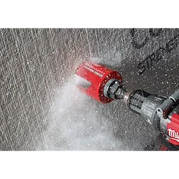 The 2-9/16" BIG HAWG™ with Carbide Teeth is shown in action, cutting through a concrete wall. The red hole saw is attached to a power drill, generating dust and debris as it operates.