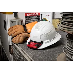 White hard hat equipped with a BOLT Personal Voltage Detector, placed on a workbench alongside a pair of brown work gloves.