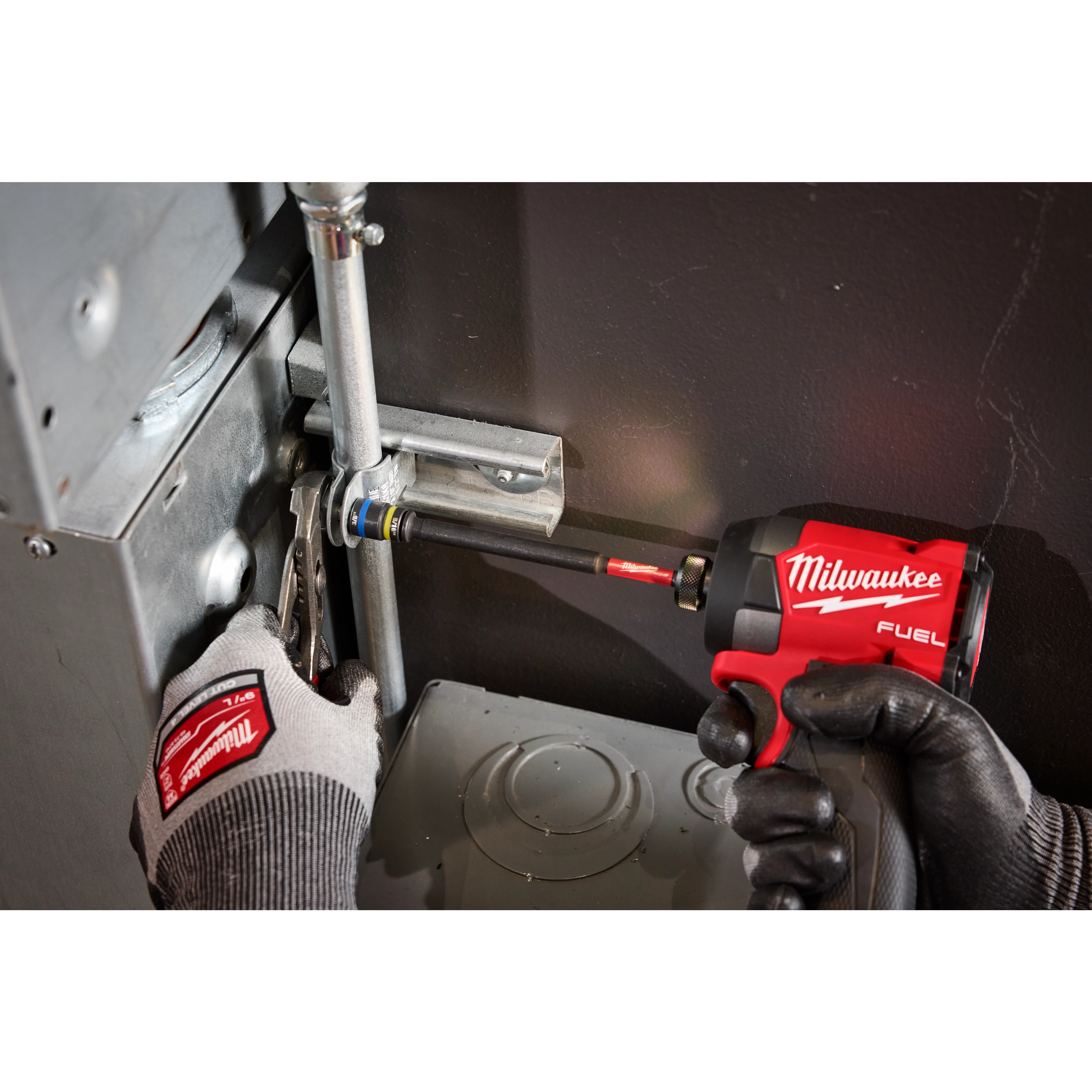 A person is using the SHOCKWAVE™ Impact Duty 5/16” and 3/8” x 6” QUIK-CLEAR™ 2-in-1 Magnetic Nut Driver attached to a red Milwaukee Fuel power tool to tighten a pipe fitting. The individual is wearing Milwaukee-branded work gloves. The setting is an industrial environment.