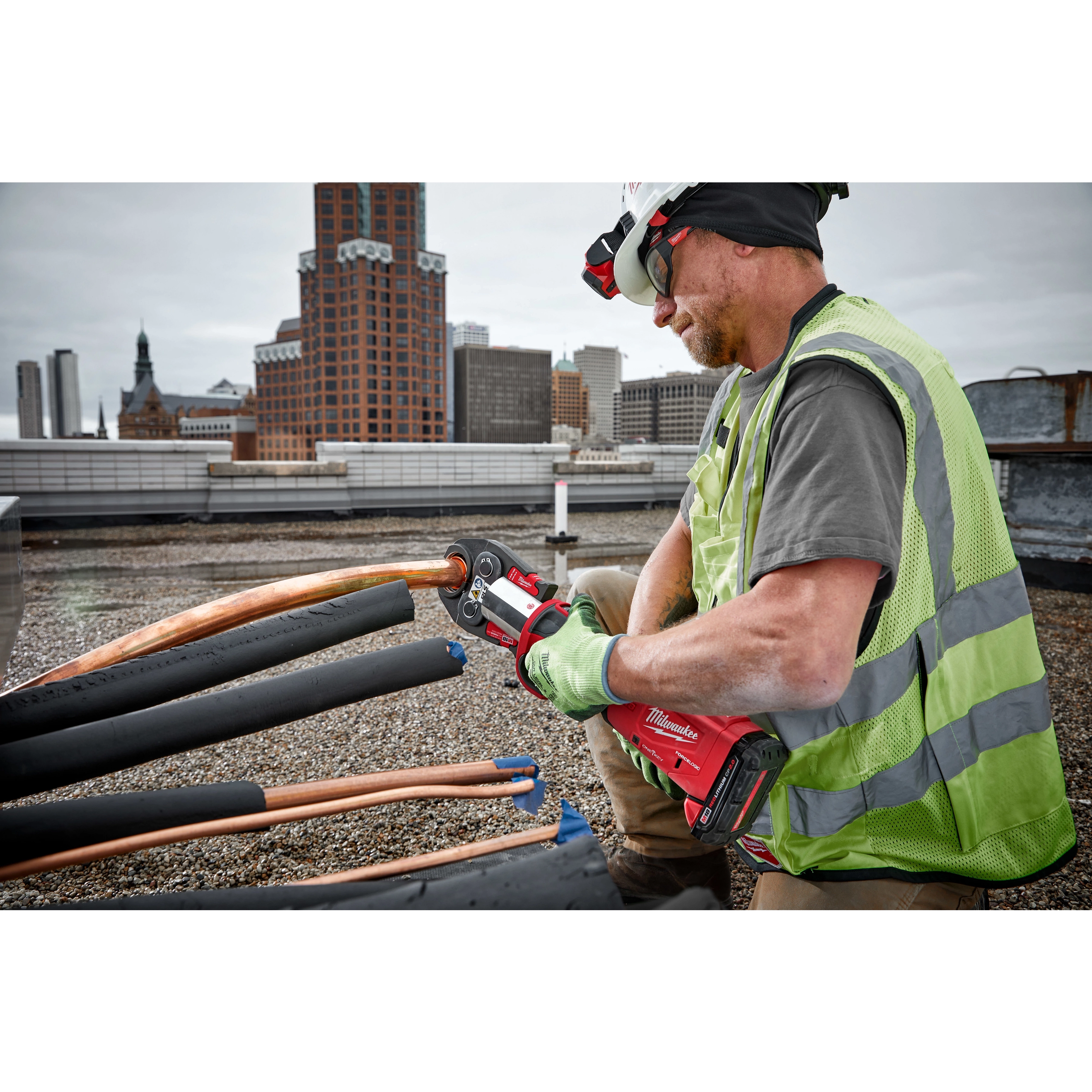 A worker uses RLS ACR Press Jaws for M18 FORCE LOGIC Press Tools on copper pipes, with a cityscape in the background.