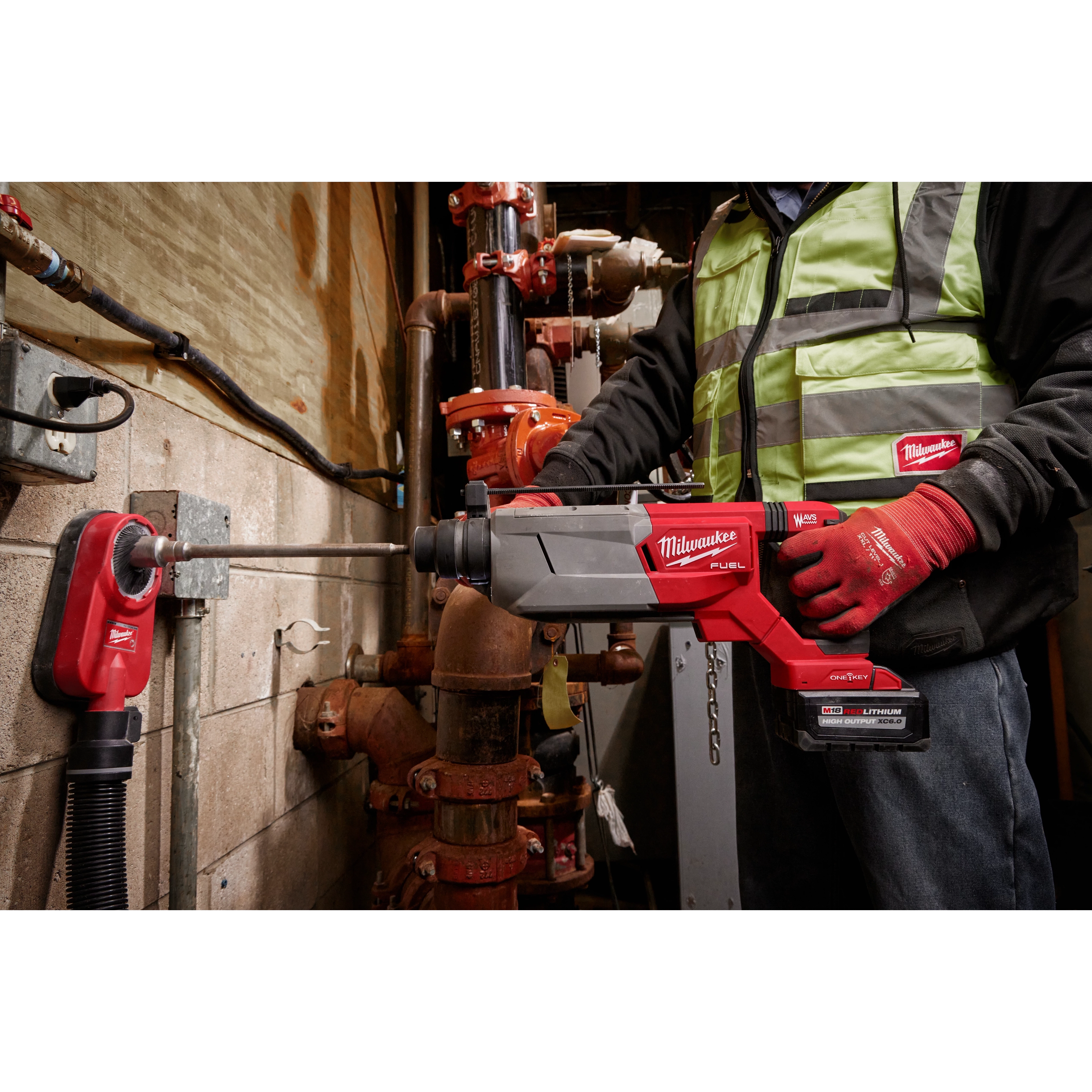 Person using the M18 FUEL 1-1/4" SDS Plus D-Handle Rotary Hammer with ONE-KEY on a concrete wall with red plumbing in the background.