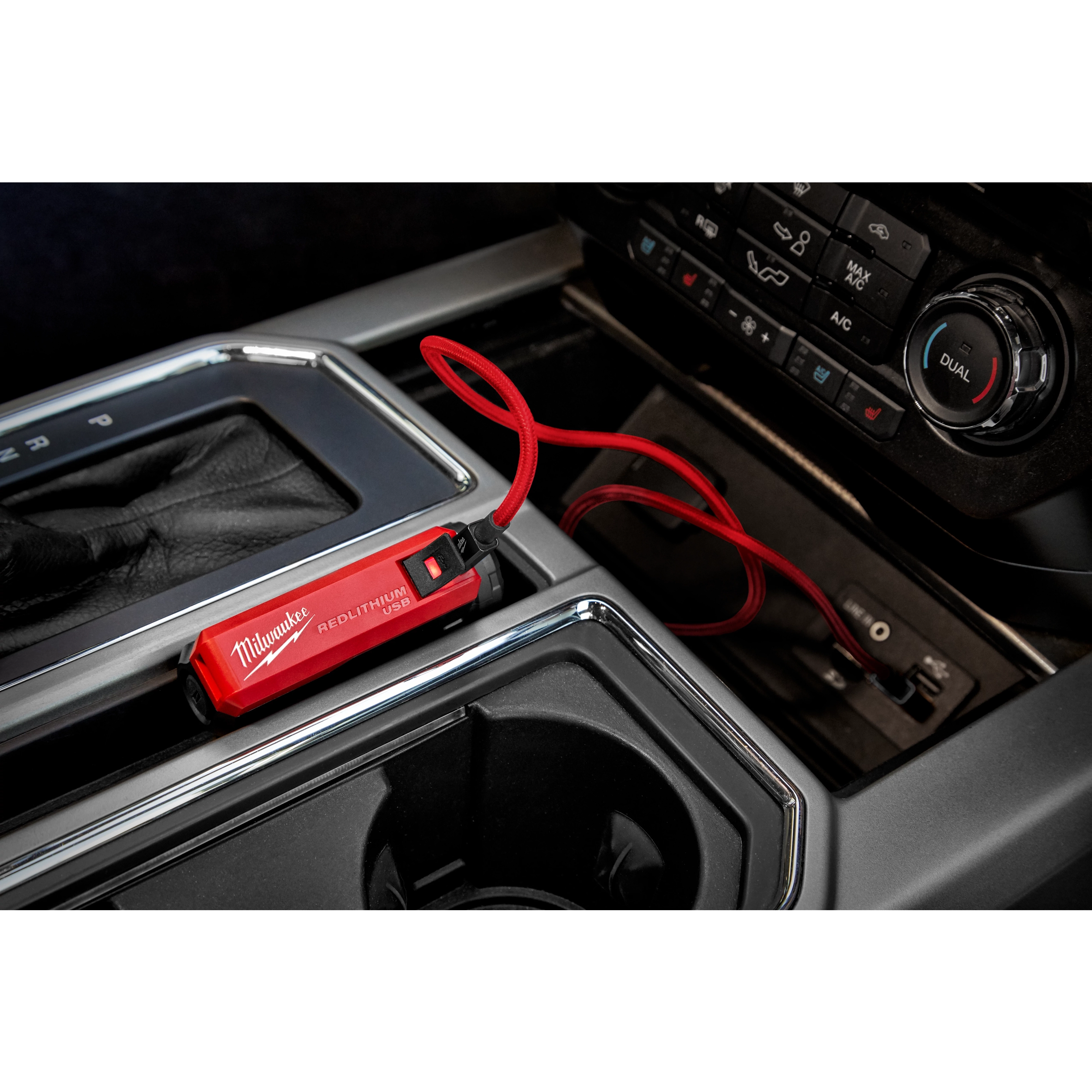 Milwaukee REDLITHIUM™ USB Charger and Portable Power Source Kit, with a red design, is connected to a USB port inside a car. The compact charger is placed near the gear shift with a red cable attached, highlighting its ease of use for charging devices on the go.