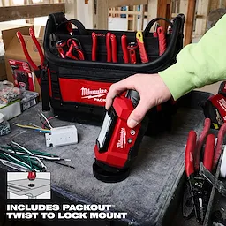 A person holds the Milwaukee M12™ ROVER™ Multi-Directional Flood Light with PACKOUT™ Compatibility. The light is placed on a workbench next to various tools and a Milwaukee tool bag. The image highlights the PACKOUT™ Twist to Lock Mount feature.