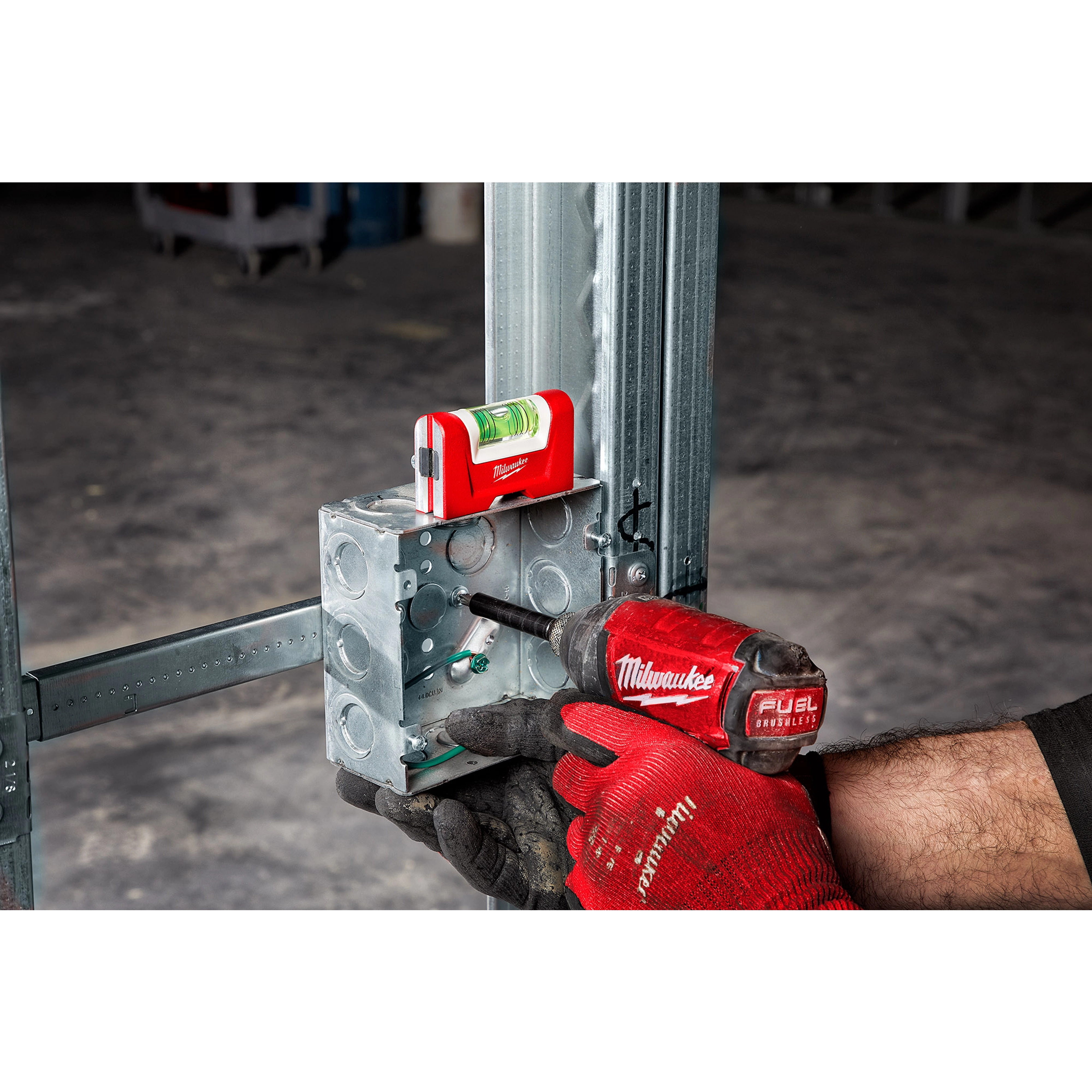 Worker using a red power drill to secure a metal junction box to a steel frame, with a Pocket Level resting on top.