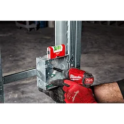 Worker using a red power drill to secure a metal junction box to a steel frame, with a Pocket Level resting on top.