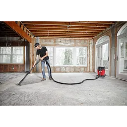 Man using a vacuum cleaner attached to a 1-7/8” x 16' flexible hose in an unfinished room with exposed insulation and large windows.