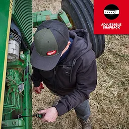 Person working on farm equipment while wearing a GRIDIRON Snapback Trucker Hat outdoors.