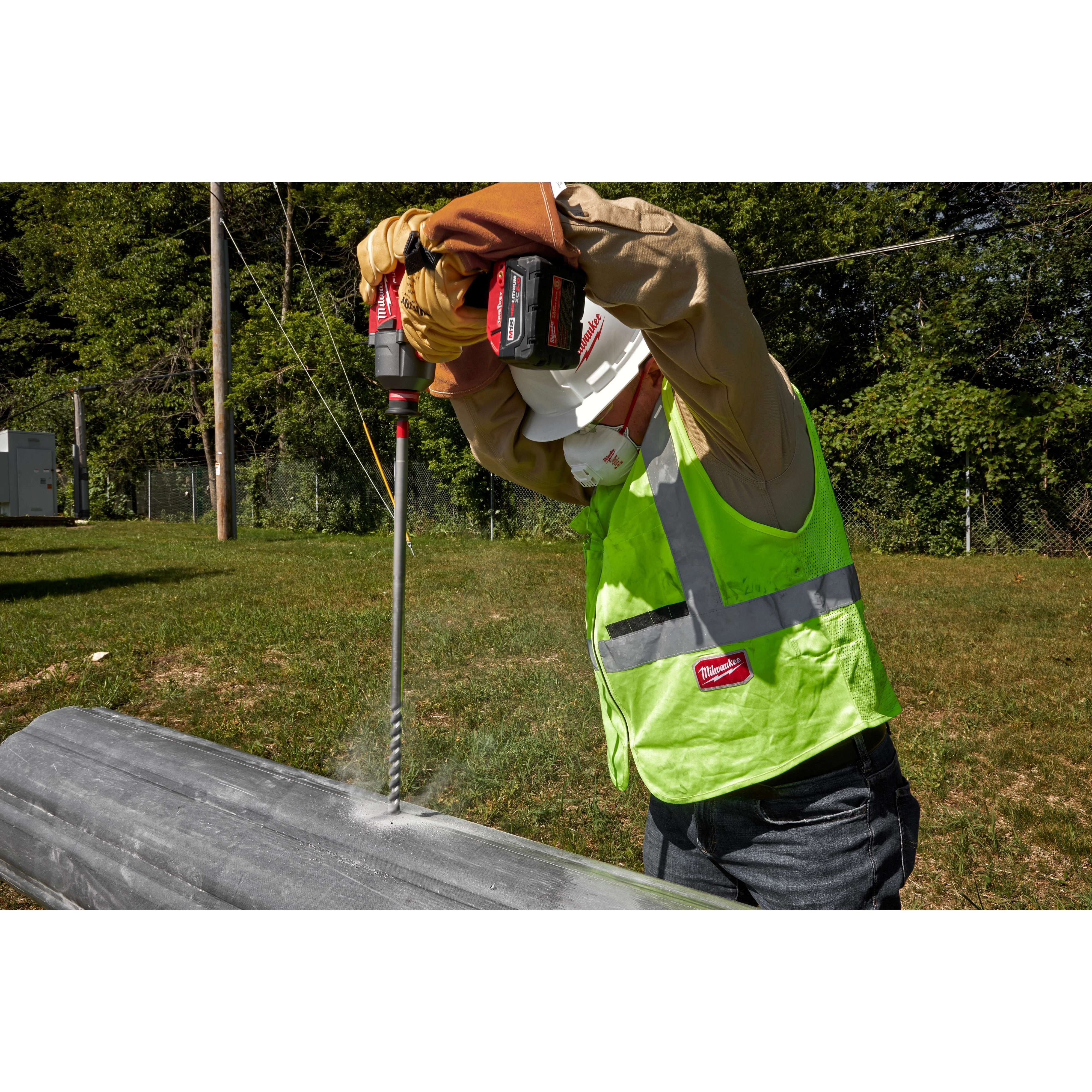 A worker in safety gear uses the 13/16” x 22” Shockwave Lineman’s Fiberglass Drill Bit to drill into a utility pole. The worker is outdoors, wearing a hard hat, gloves, high-visibility vest, and a long-sleeve shirt. The drill is cordless with a red and black design.