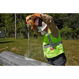 A worker in safety gear uses the 13/16” x 22” Shockwave Lineman’s Fiberglass Drill Bit to drill into a utility pole. The worker is outdoors, wearing a hard hat, gloves, high-visibility vest, and a long-sleeve shirt. The drill is cordless with a red and black design.
