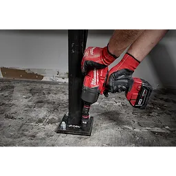 A person uses a red power tool equipped with a SHOCKWAVE™ Impact Duty™ 1/2" Drive 15/16" Deep 6 Point Socket to secure a bolt on a metal base. The tool is being held with gloved hands on a concrete floor.