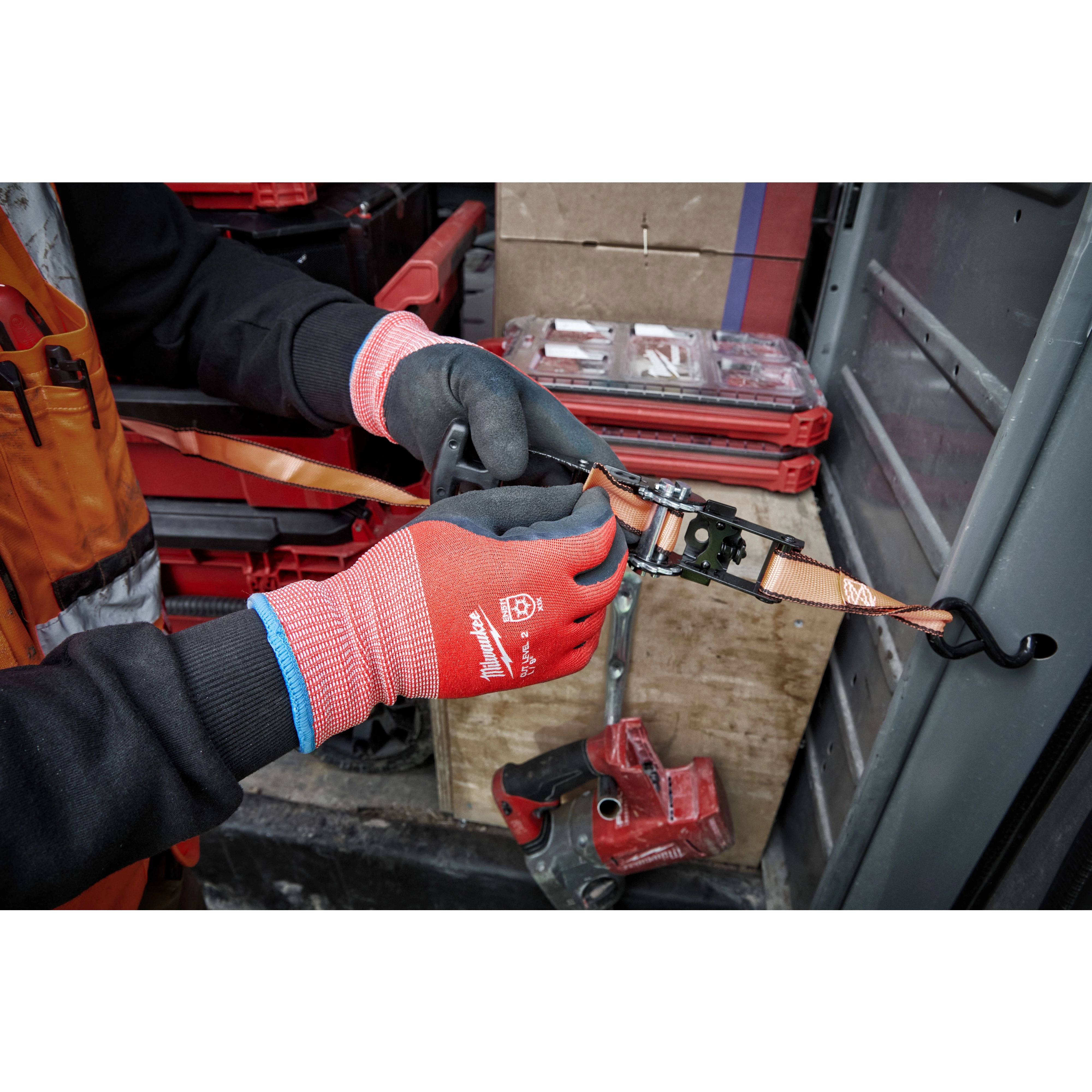 Person wearing Cut Level 2 Winter Dipped Gloves - L, adjusting a ratchet strap inside a workshop with toolboxes and equipment nearby.