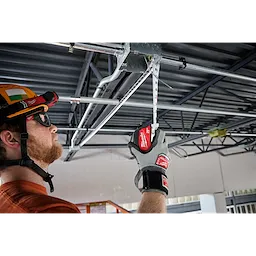 A person wearing gloves is using a 25ft Electrician's Magnetic Tape Measure to measure piping attached to a metal beam on a ceiling. The tape measure is extended and held in place, and the person is positioned on the left side of the image.