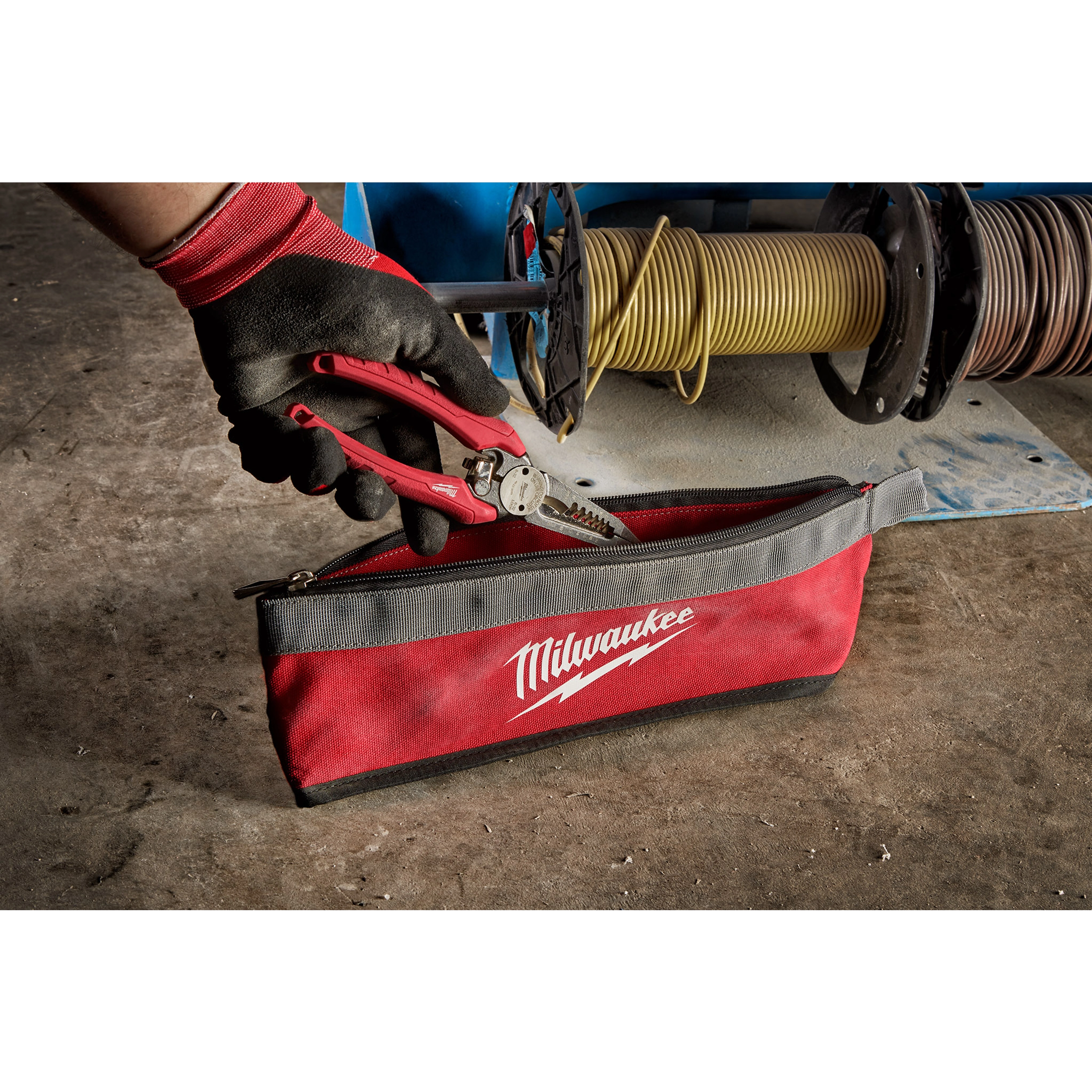 A gloved hand holding pliers above a red and gray Milwaukee Zipper Pouch on a workshop floor, with cables in the background.