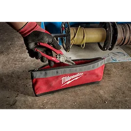A gloved hand holding pliers above a red and gray Milwaukee Zipper Pouch on a workshop floor, with cables in the background.