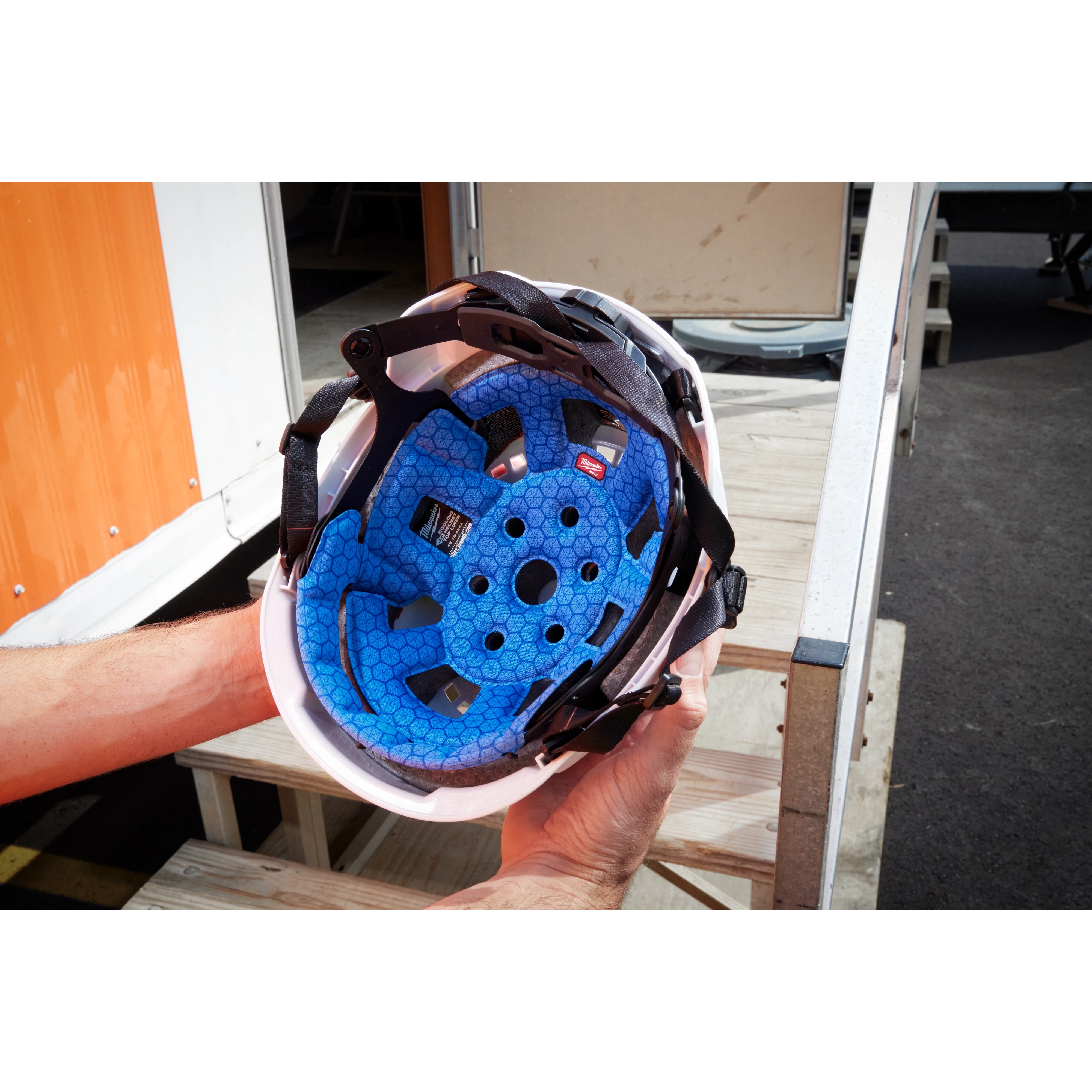 A pair of hands holds a white helmet featuring the BOLT™ Safety Helmet Cooling Sweat Band and Liner, along with the BOLT™ Safety Helmet Cooling Liner in blue, designed for enhanced comfort and cooling. The background shows outdoor steps and part of an orange and white building.