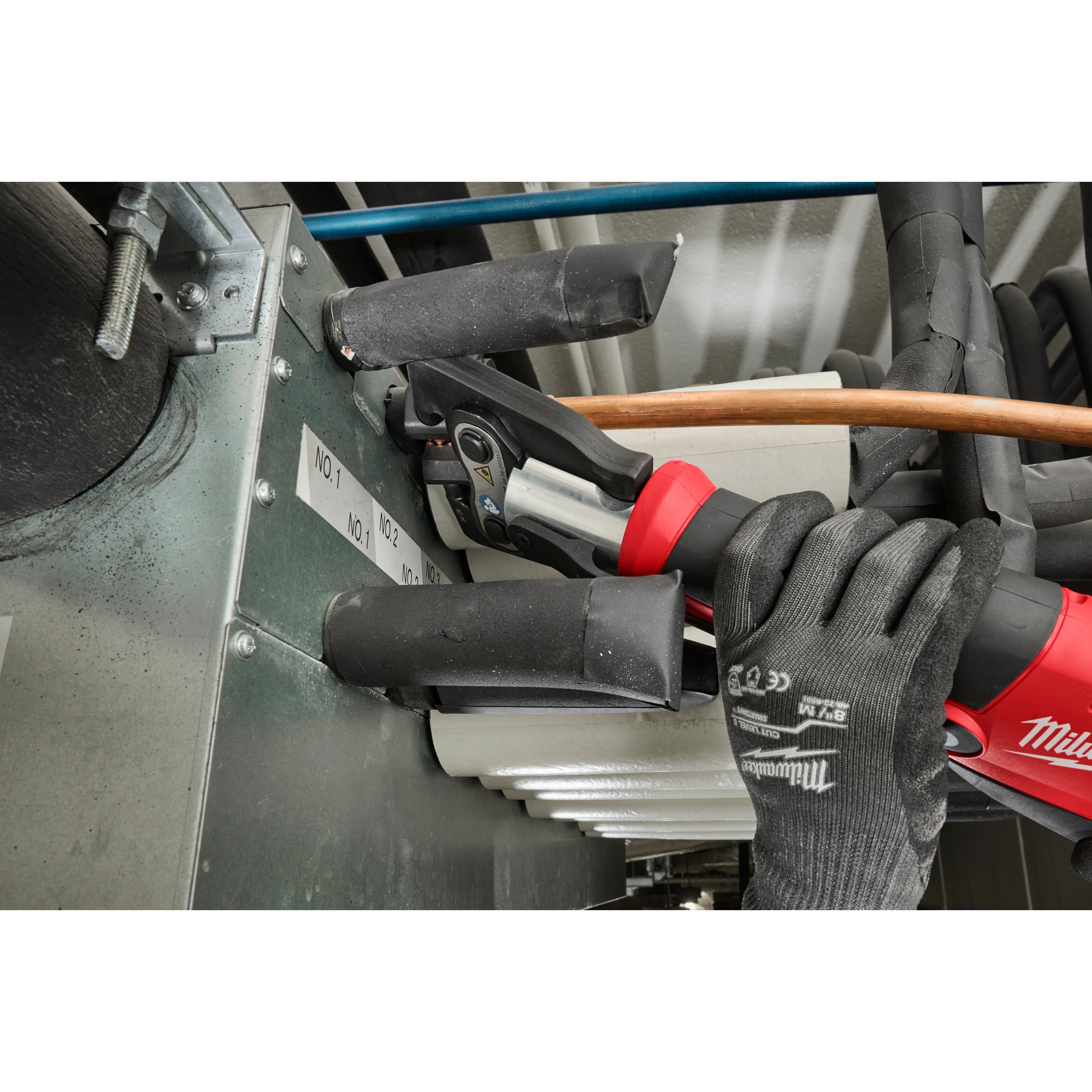 A worker uses the 1/4" - 7/8" Streamline® ACR Pivoting Press Ring Kit for M18™ FORCE LOGIC™ Press Tool to install copper piping in a metallic unit. The worker wears black gloves and operates the red and black press tool, which is clamped onto the copper pipe.
