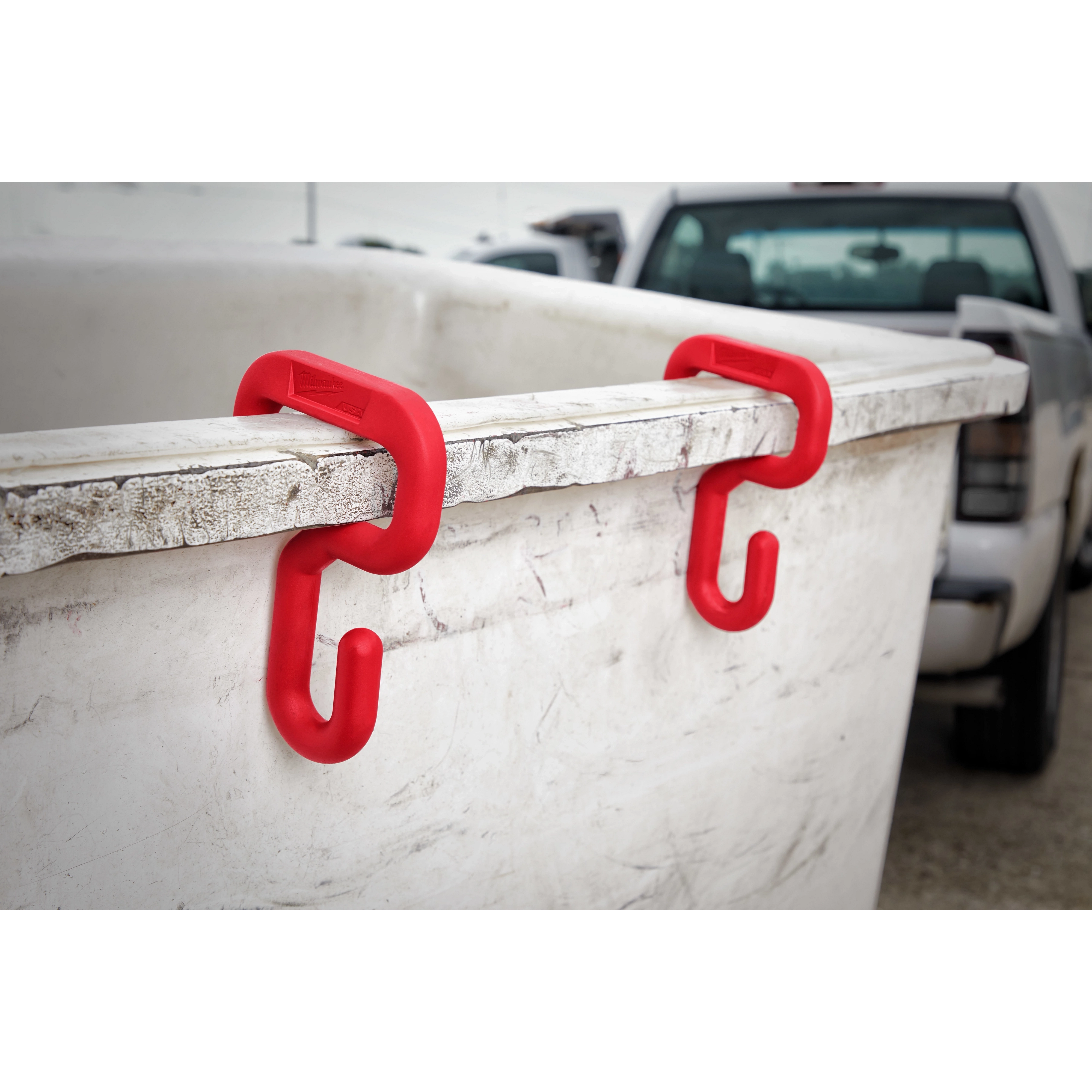 Red plastic hooks labeled "3%22%20Bucket%20Hook" are attached to the rim of a large white bin. A pickup truck is in the background.