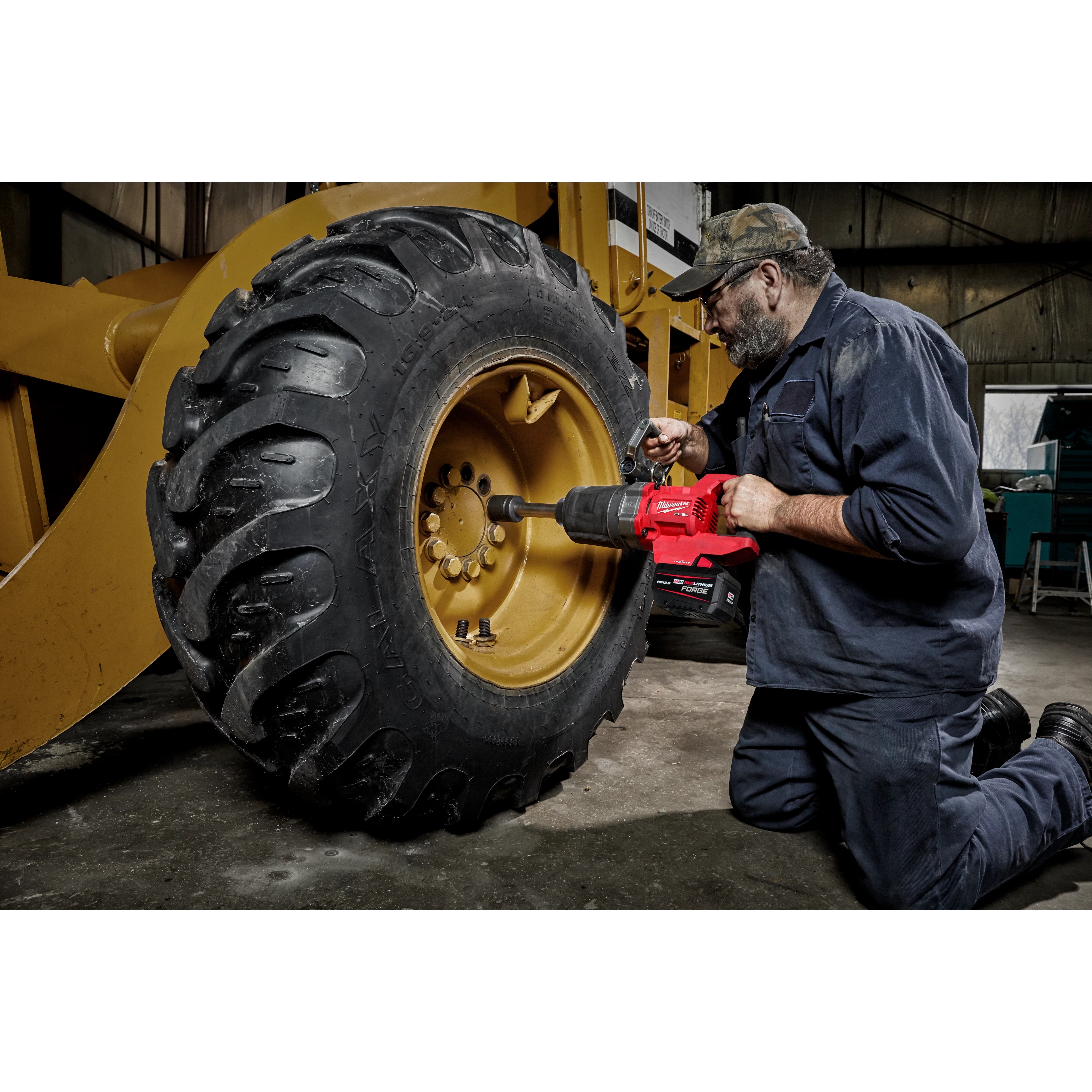 A person uses an M18 FUEL™ 1" D-Handle Ext. Anvil High Torque Impact Wrench with ONE-KEY™ App Shot to work on the wheel of a large, yellow construction vehicle. The person is kneeling on the ground while operating the tool.