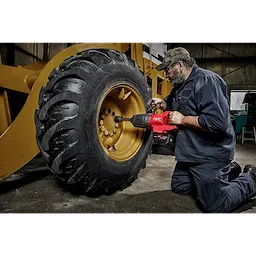 A person uses an M18 FUEL™ 1" D-Handle Ext. Anvil High Torque Impact Wrench with ONE-KEY™ App Shot to work on the wheel of a large, yellow construction vehicle. The person is kneeling on the ground while operating the tool.