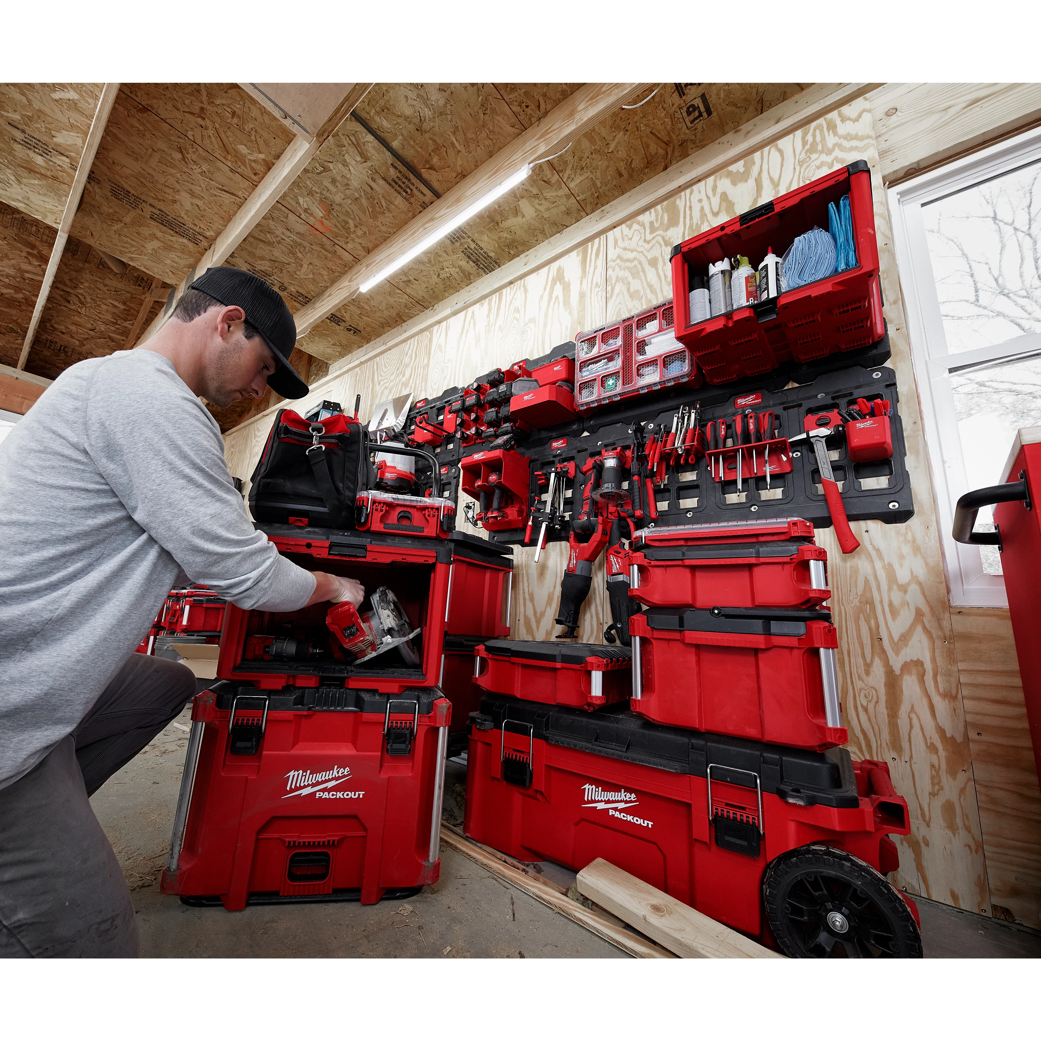 A person uses a PACKOUT™ Cabinet, which is part of a red modular storage system filled with various tools. The setup is mounted on a wooden wall within a work shed, with additional storage units organized efficiently on and around the cabinet.