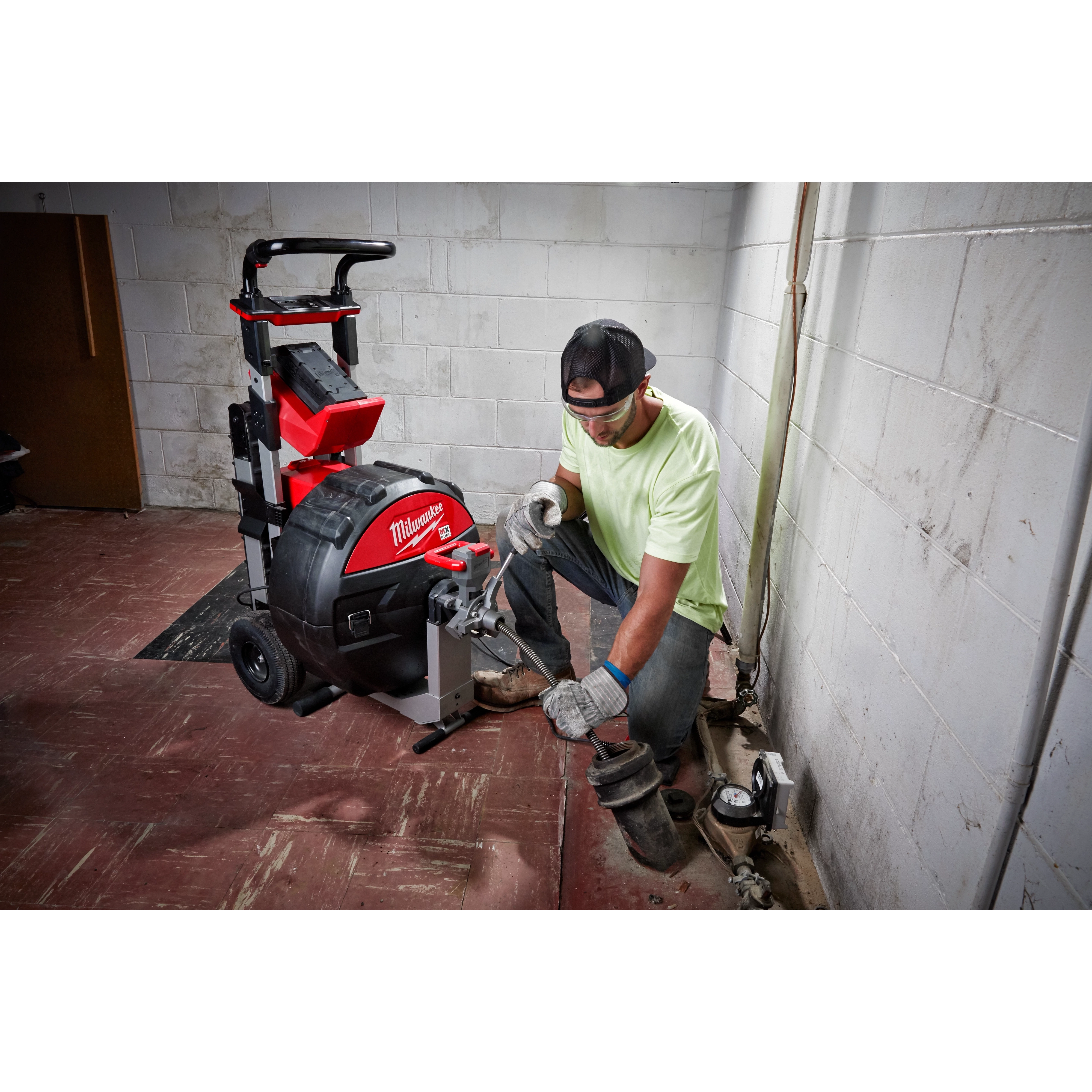 A person crouches beside a Milwaukee-branded MX FUEL™ Sewer Drum Machine w/ POWERTREDZ™ in a utility room with white tiled walls. The machine has a red and black body and is equipped with large wheels and a handle for mobility.