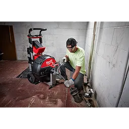 A person crouches beside a Milwaukee-branded MX FUEL™ Sewer Drum Machine w/ POWERTREDZ™ in a utility room with white tiled walls. The machine has a red and black body and is equipped with large wheels and a handle for mobility.
