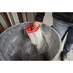 Person cleaning a Milwaukee Large Wet/Dry Vacuum HEPA Filter by tapping it over a trash can in a workshop.