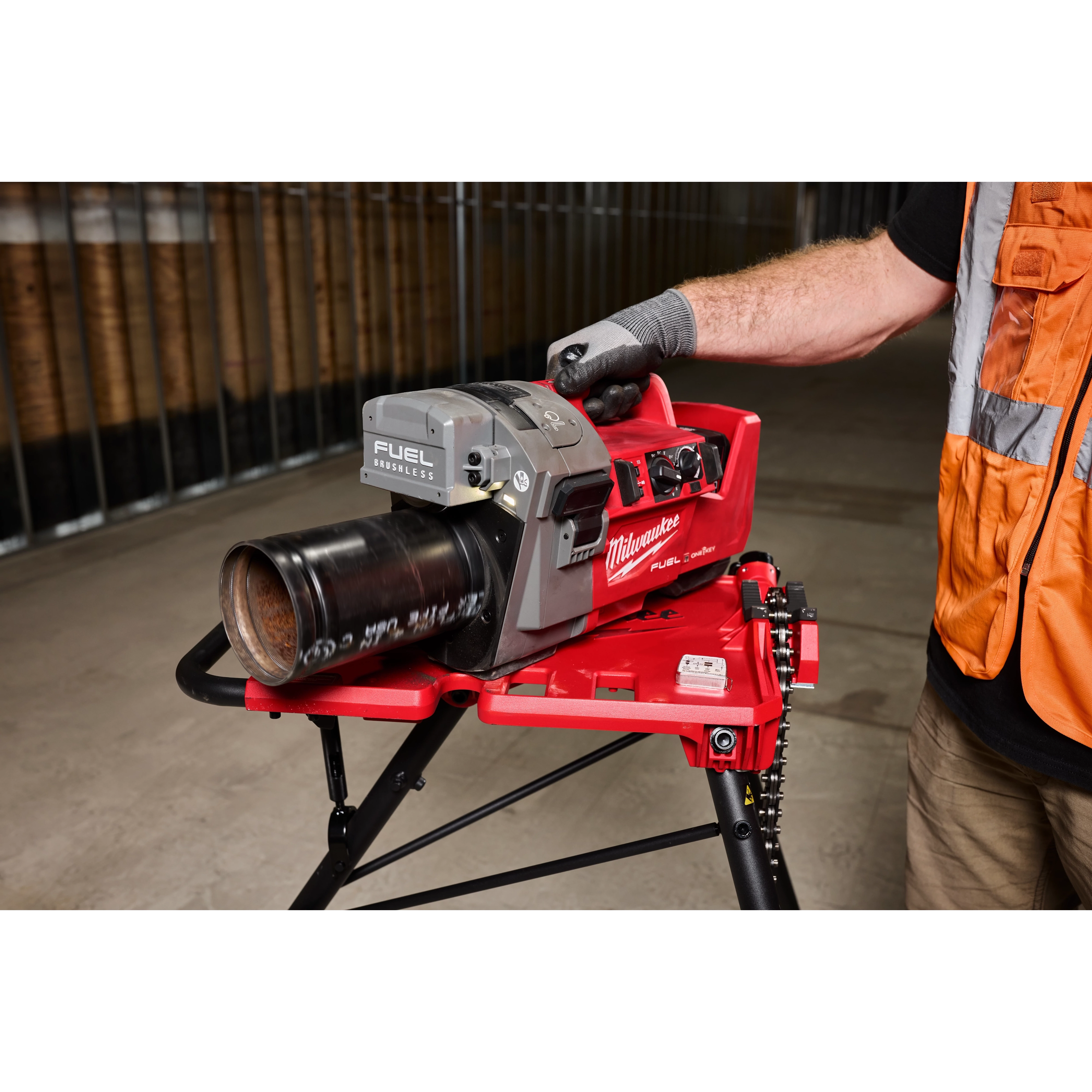 A person operating an M18 FUEL™ RINGER™ Roll Groover for 1-1/4” - 4” Sch 10/40, 6” Sch 10. The red and gray tool is mounted on a stand, with a gloved hand guiding a pipe through the machine. The background shows an industrial setting.