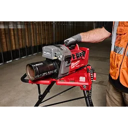 A person operating an M18 FUEL™ RINGER™ Roll Groover for 1-1/4” - 4” Sch 10/40, 6” Sch 10. The red and gray tool is mounted on a stand, with a gloved hand guiding a pipe through the machine. The background shows an industrial setting.