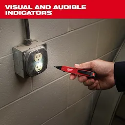 A hand holds a red and black Milwaukee voltage detector next to an electrical outlet on a gray wall. The device lights up, indicating the presence of voltage. The image includes text "VISUAL AND AUDIBLE INDICATORS" in bold white font above the product.