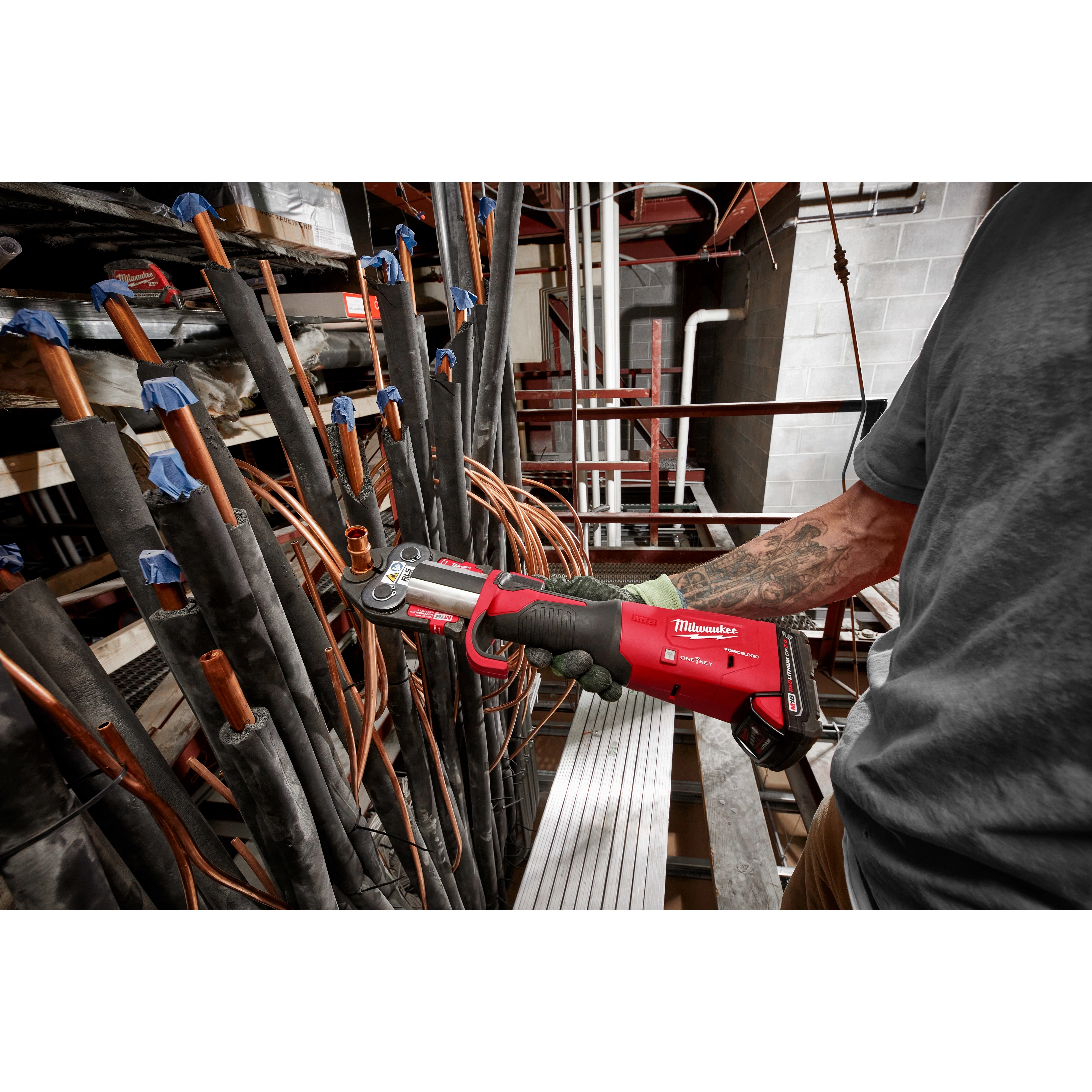 A person operates RLS ACR Press Jaws for M18 FORCE LOGIC Press Tools on a copper pipe in an industrial setting with various pipes.