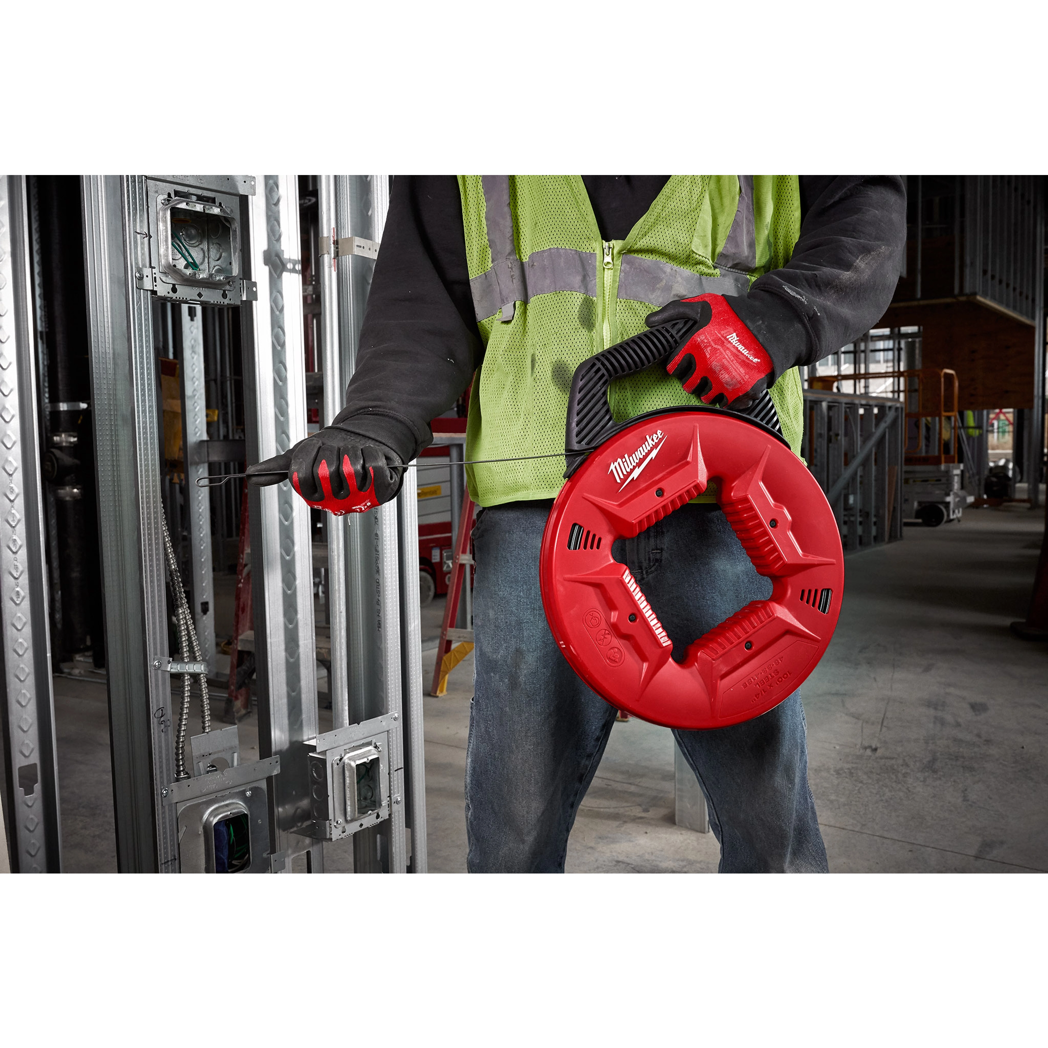 Construction worker uses 1/4" 100' Steel Fish Tape with a red case and black handle, wearing safety gear on a job site.