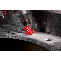 A Milwaukee® 1-3/8" DIAMOND MAX™ Hole Saw Bit attached to a drill is cutting into a grey stone surface, creating a hole. Dust and debris are visible around the drilling area.