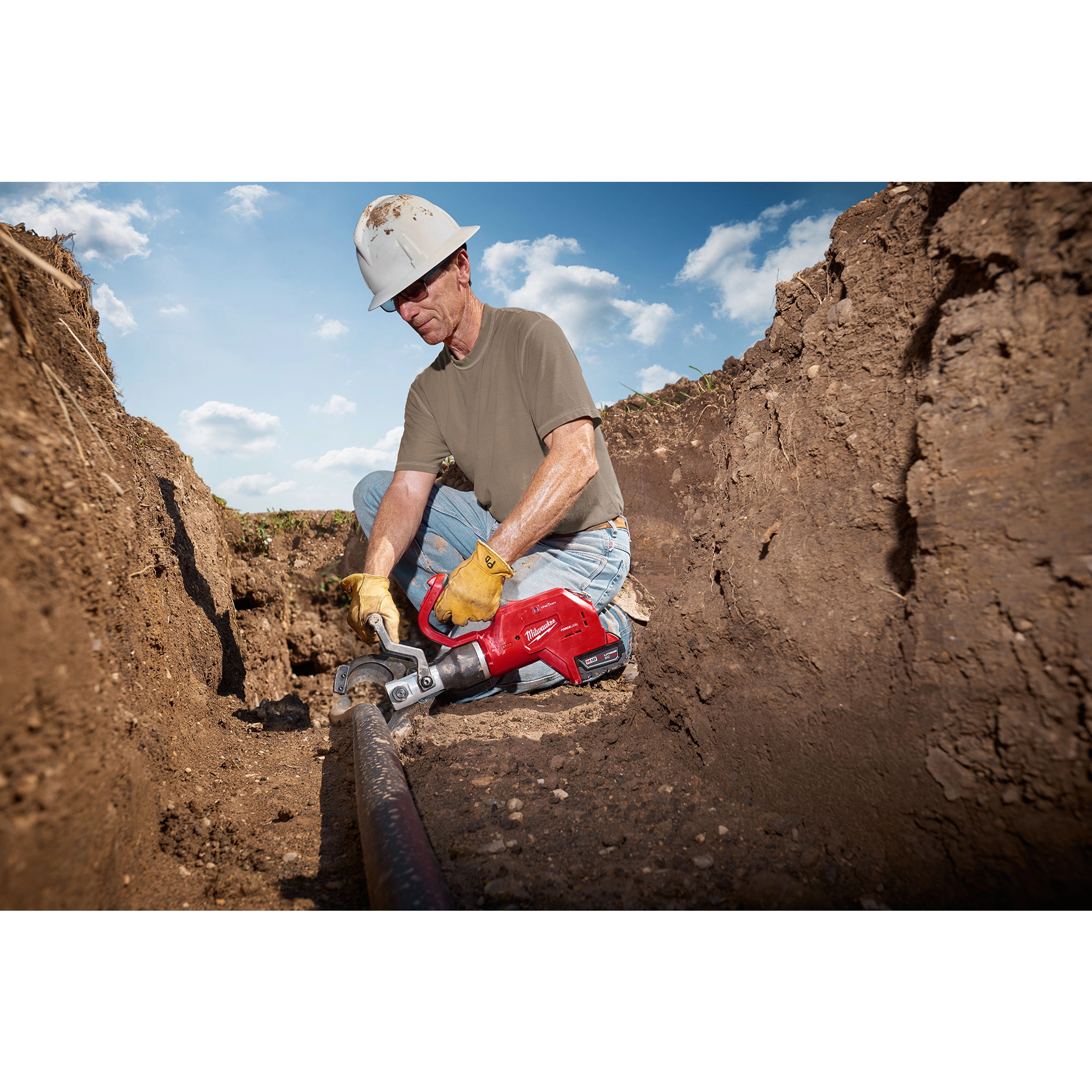 A person kneels in a trench using the FORCELOGIC™ M18™ 3" Underground Cable Cutter Kit to cut a cable. The red cable cutter is held with both hands and is cutting through a thick underground cable. The kit appears durable and suitable for heavy-duty tasks.