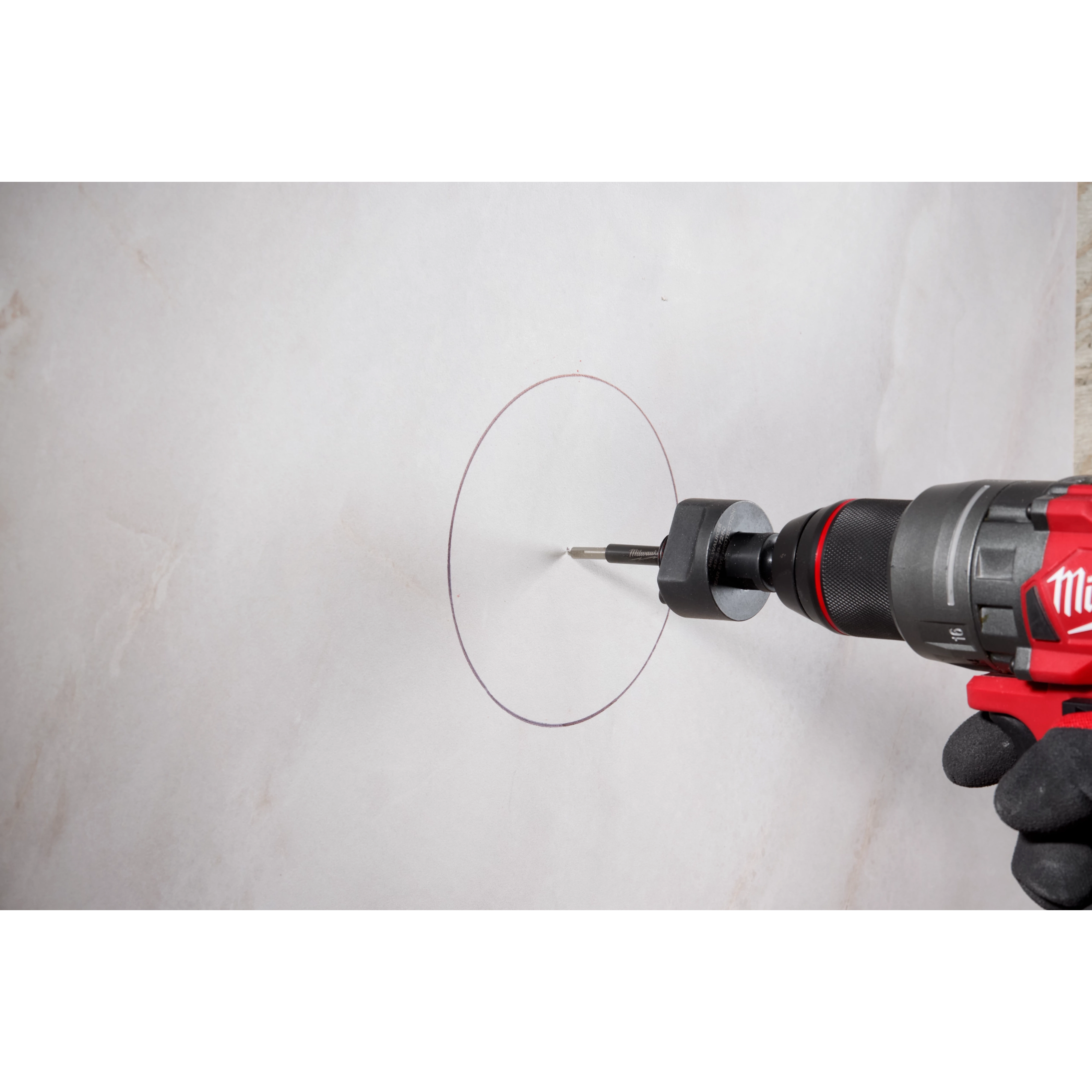 A Milwaukee® Arbor with a Retractable Starter Bit is attached to a red and black power drill. The drill and bit are positioned against a marked circular outline on a light-colored surface, presumably preparing to cut through it.