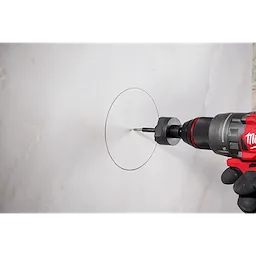 A Milwaukee® Arbor with a Retractable Starter Bit is attached to a red and black power drill. The drill and bit are positioned against a marked circular outline on a light-colored surface, presumably preparing to cut through it.