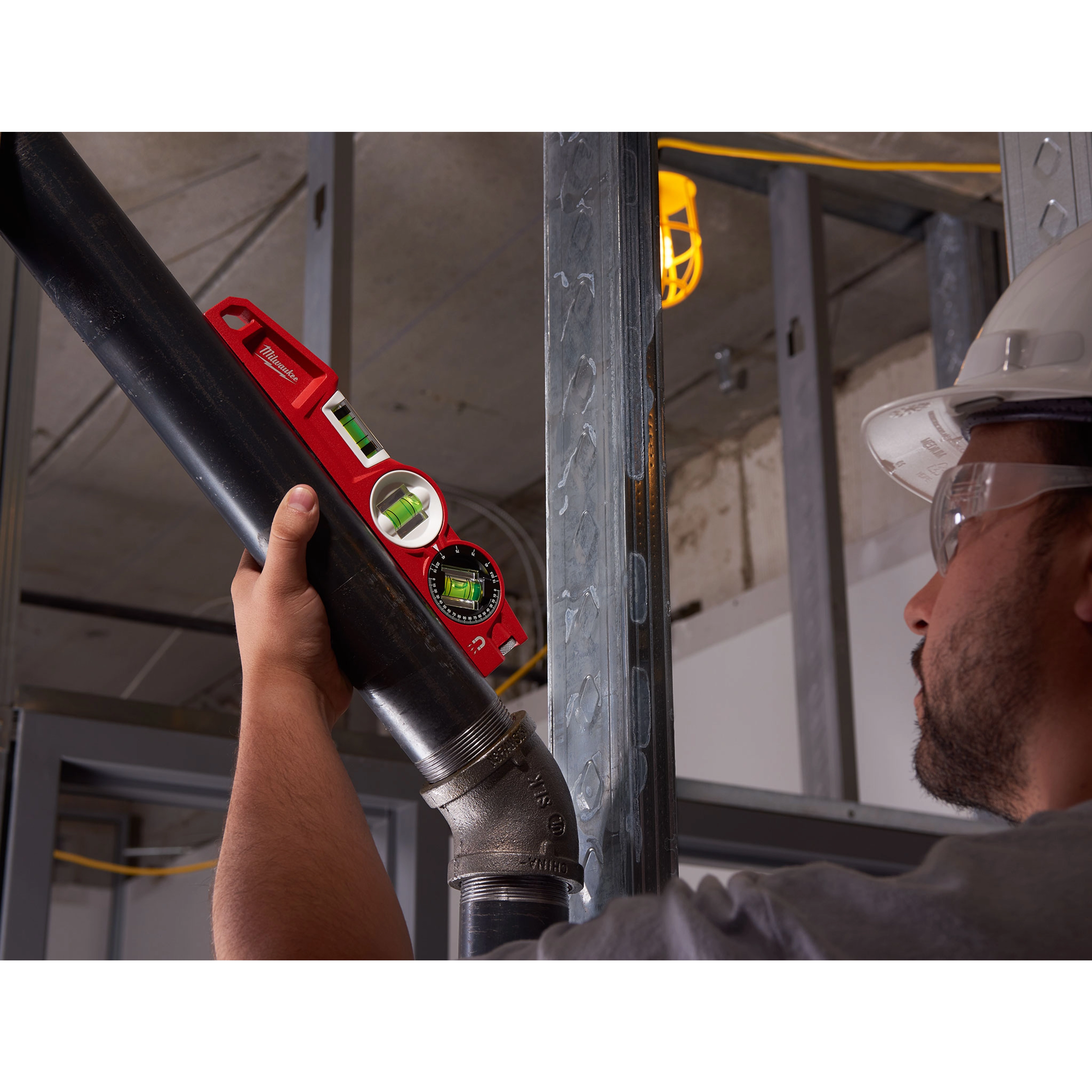 A person installs a pipe using a Block Torpedo Level in a construction setting. The level is red with two green bubble vials.