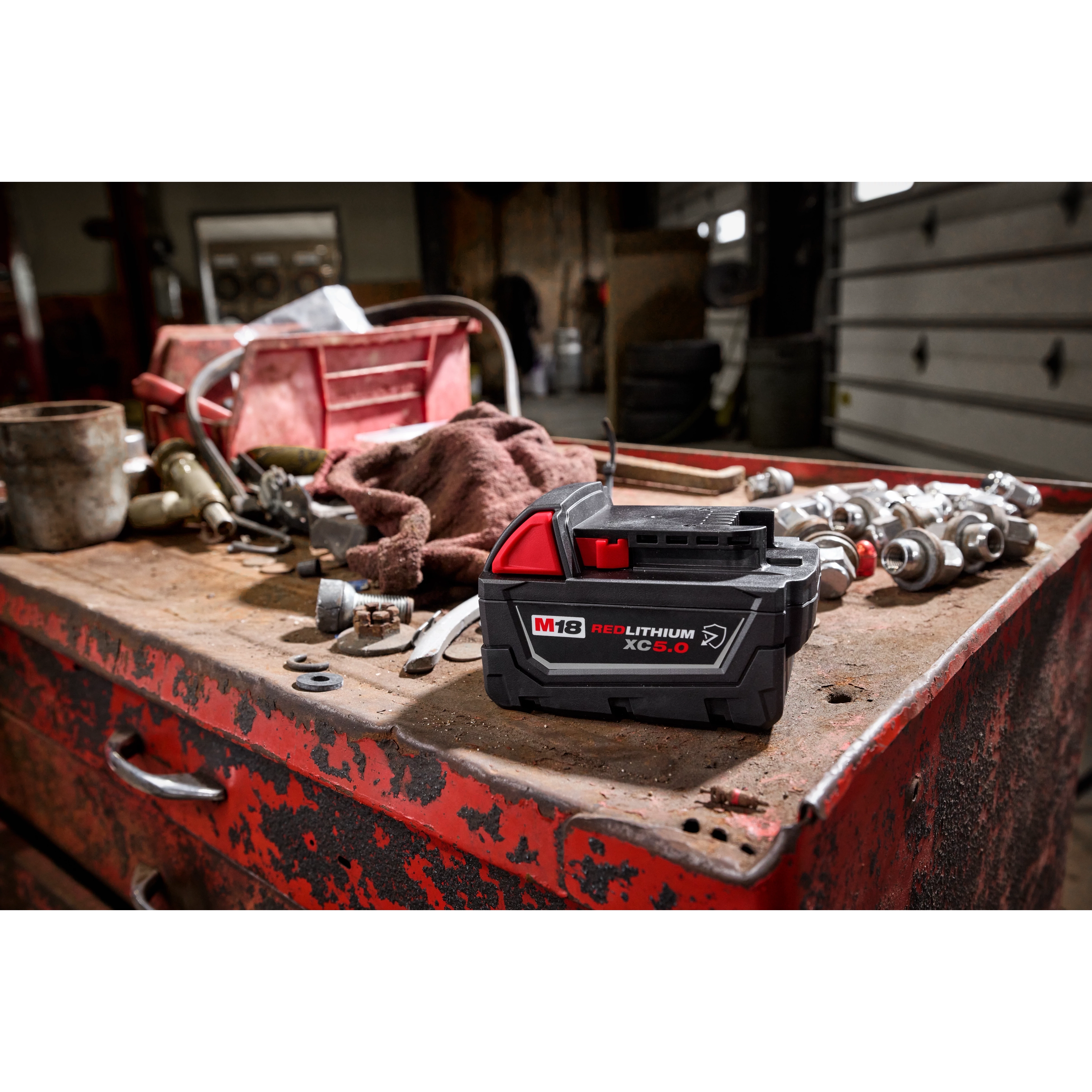 M18 REDLITHIUM XC5.0 Resistant Battery on a red, weathered workbench with various tools and fittings in a workshop setting.
