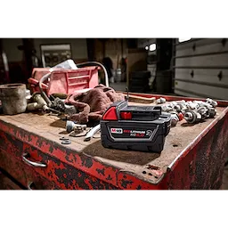 M18 REDLITHIUM XC5.0 Resistant Battery on a red, weathered workbench with various tools and fittings in a workshop setting.