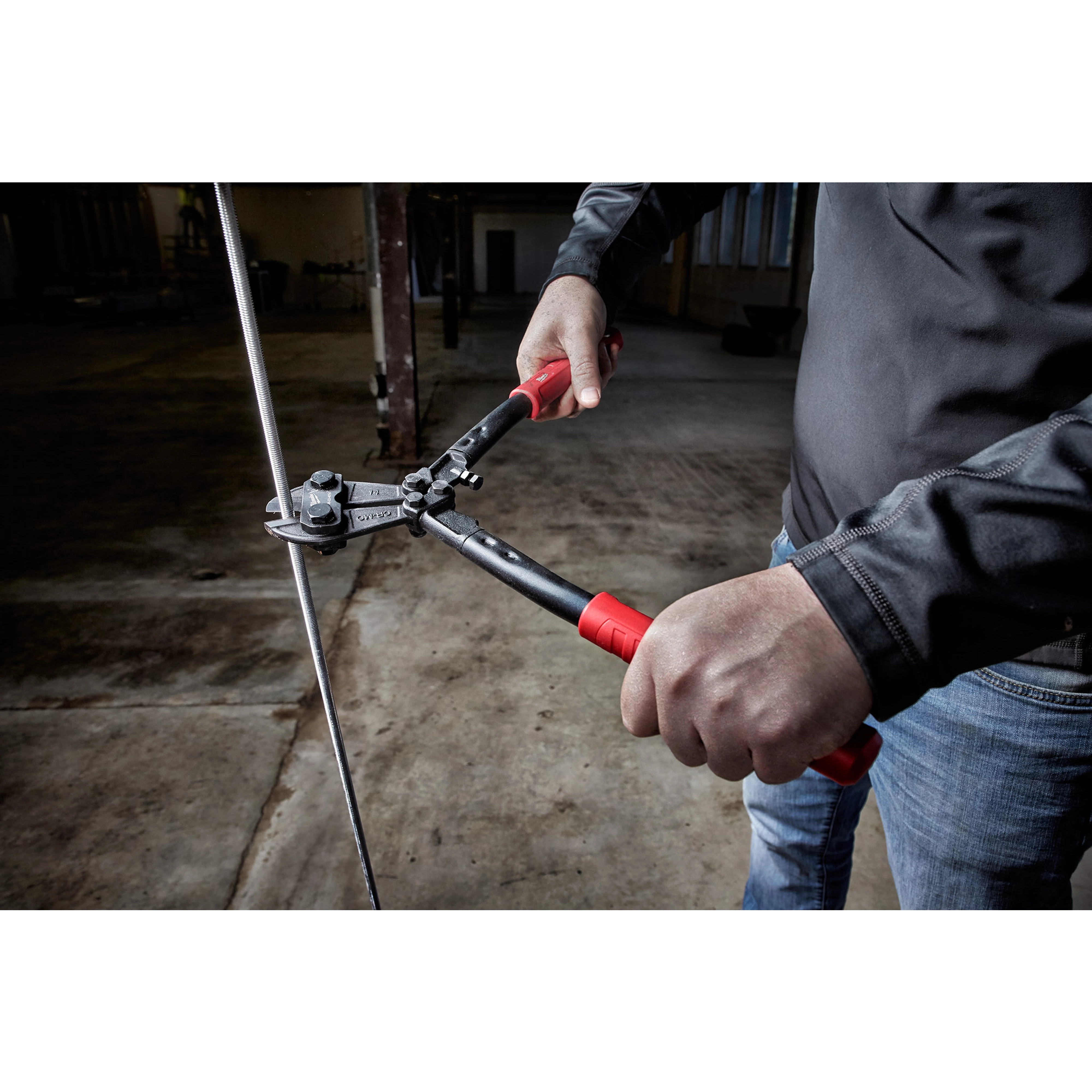 Person using 14" Bolt Cutter with red and black handles to cut through metal wire in a dimly lit, industrial setting.