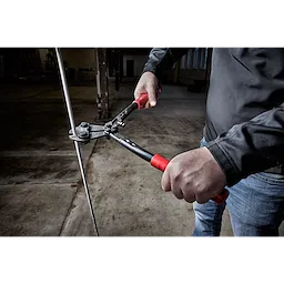 Person using 14" Bolt Cutter with red and black handles to cut through metal wire in a dimly lit, industrial setting.