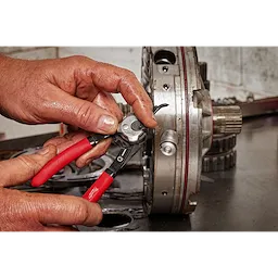 A person uses the Convertible Snap Ring Pliers Set to work on a mechanical component. The pliers have red handles, and the user’s hands are visibly dirty, indicating they are in the middle of a repair task. The mechanical component appears to be part of a larger machine or vehicle.