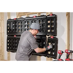 A person is mounting a PACKOUT™ Large Wall Plate on a wooden wall. Several black wall plates are already installed. Two cordless drills and a battery charger are positioned at the lower right side of the image. Other wall plates remain empty, waiting to be filled with tools.