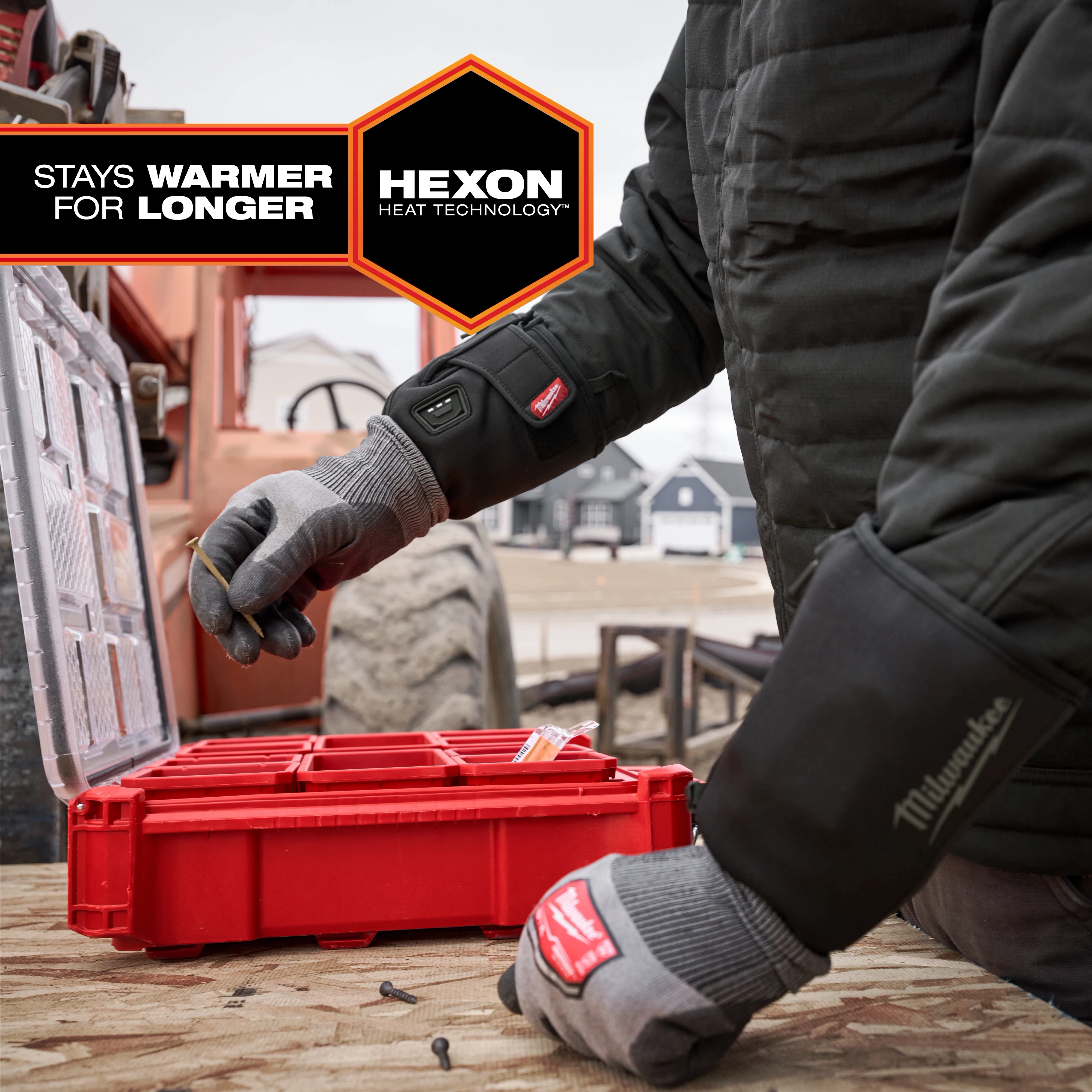 A person wearing Milwaukee Heated Work Glove Liners organizes items in a red storage box outdoors. The gloves feature Hexon Heat Technology and promise to stay warmer for longer. A construction site and large machinery are visible in the background. The gloves have black cuffs.