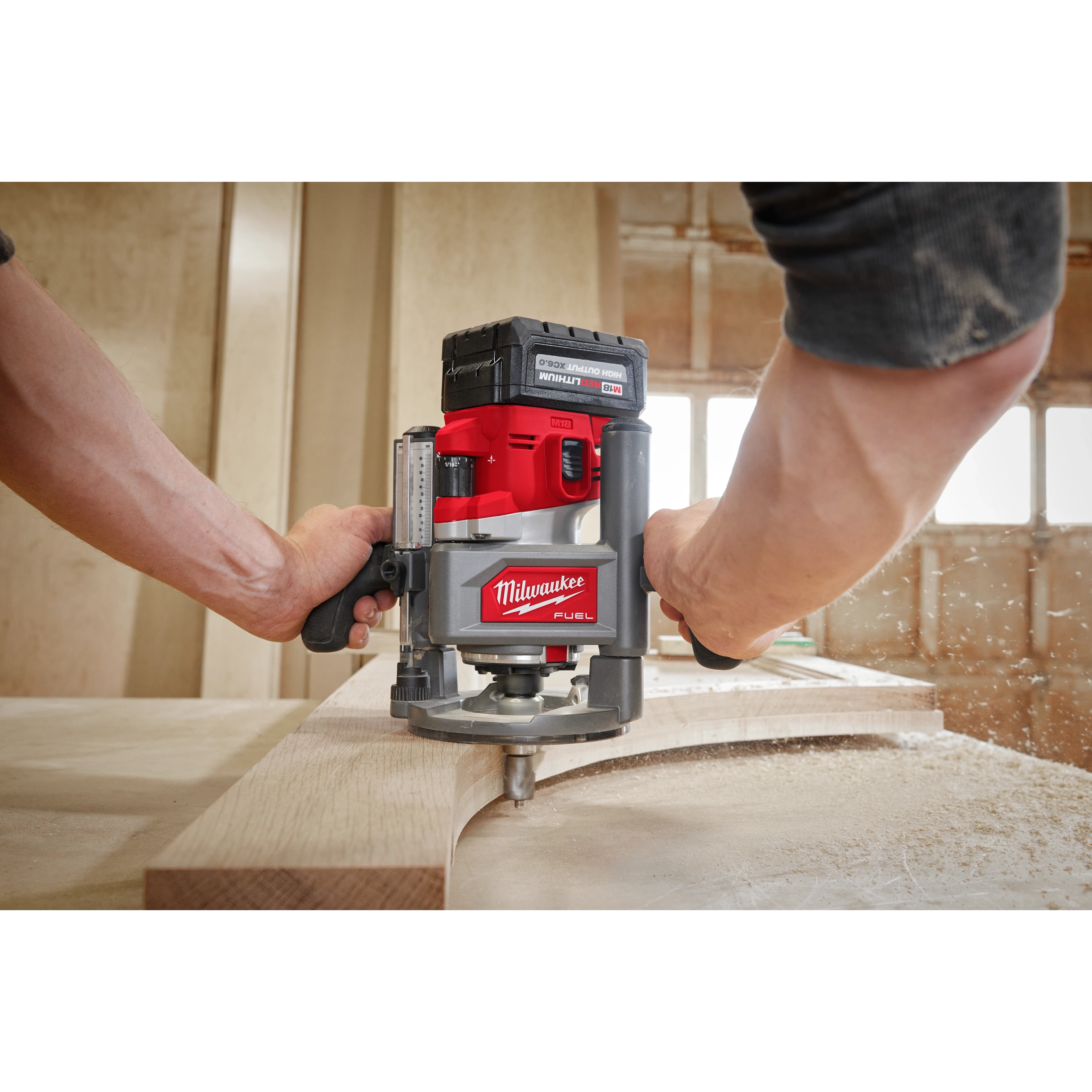 Person using a red M18 FUEL 1/2" Router to shape a piece of wood in a workshop, with sawdust flying.