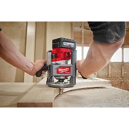 Person using a red M18 FUEL 1/2" Router to shape a piece of wood in a workshop, with sawdust flying.