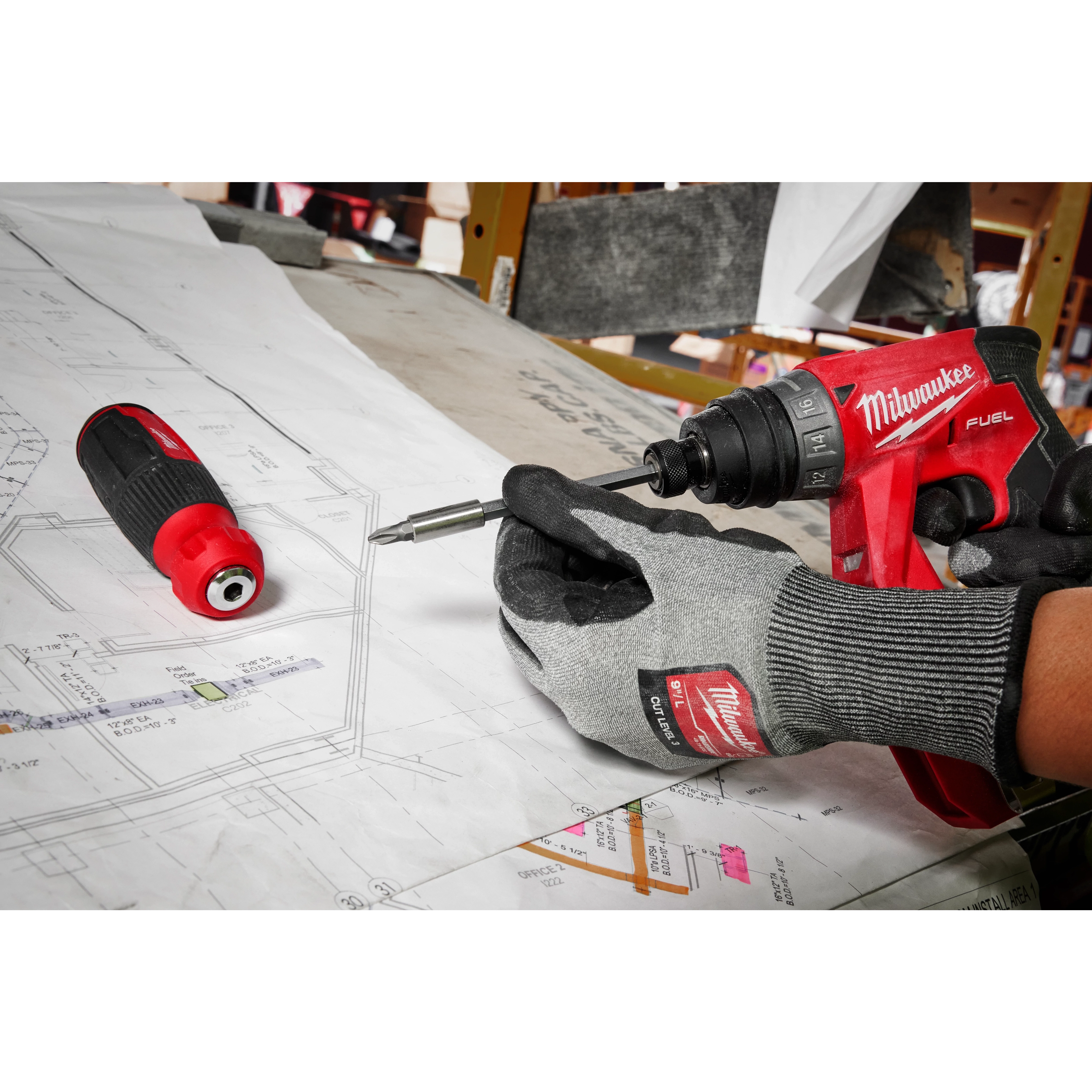 Worker using a red power drill to attach a bit to the 14-in-1 Multi-Bit Screwdriver, positioned on detailed blueprints on a desk.