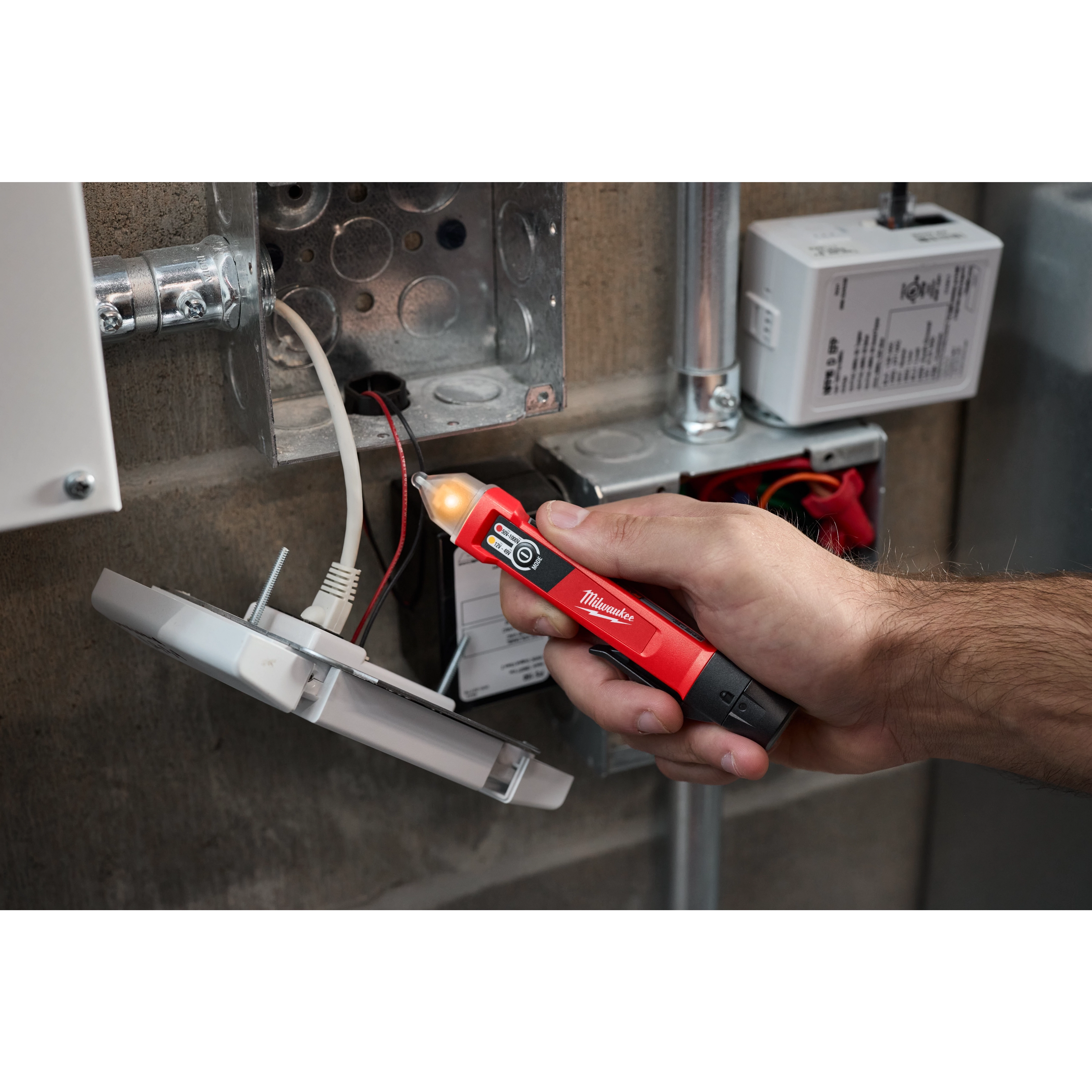 A person uses a Dual Range Voltage Detector to test wiring in an open electrical box. The red tool has a black tip and a glowing light, indicating the presence of voltage.