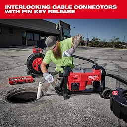 Person using Milwaukee interlocking cable connectors with pin key release system to insert a drainage tool into a large outdoor drainage pipe. Various Milwaukee tools and equipment are arranged nearby on the ground, including handheld tools and a portable reel.